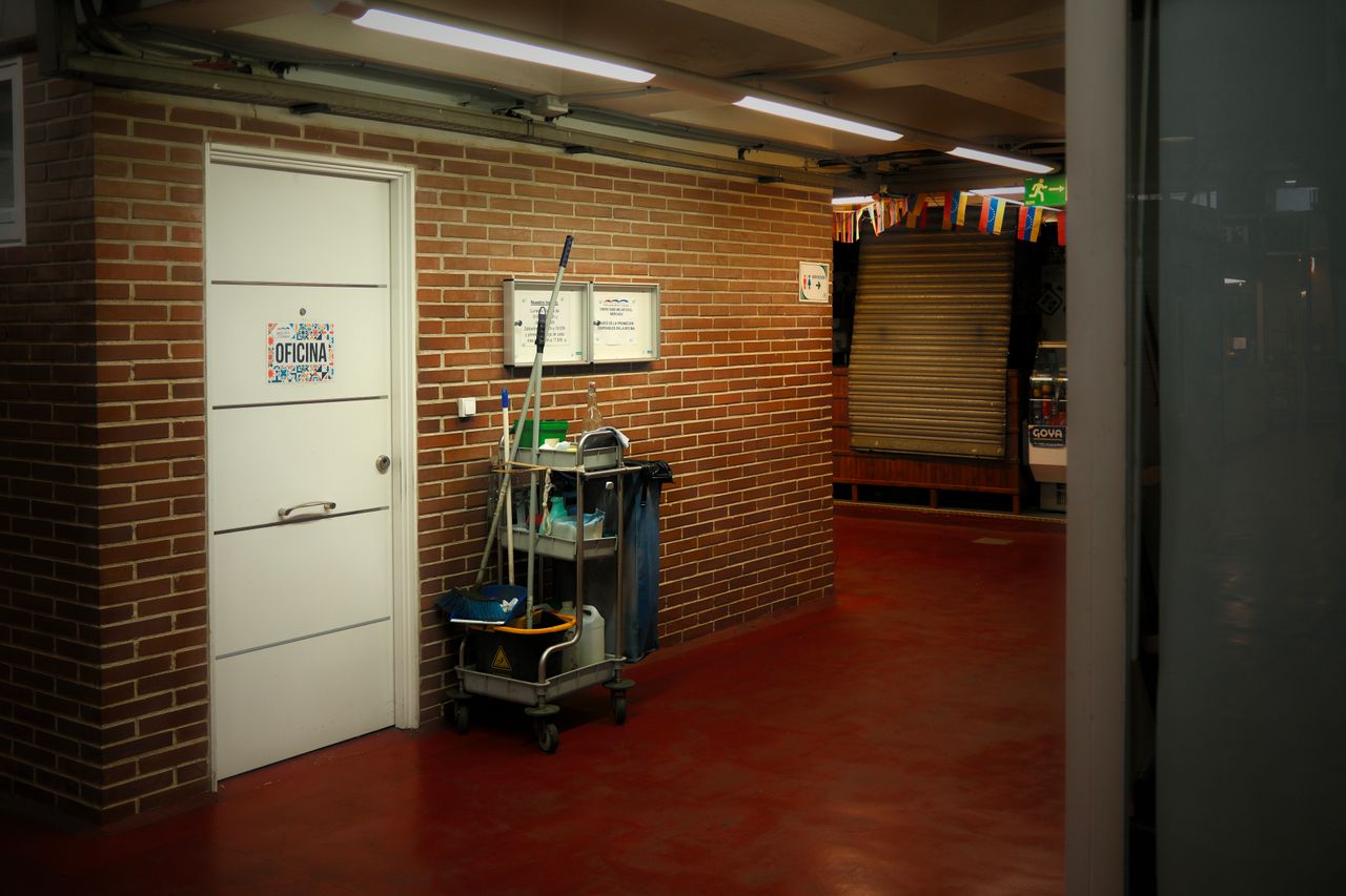 Un couloir industriel vivement éclairé, avec un chariot de nettoyage et du matériel près d’une porte de bureau.